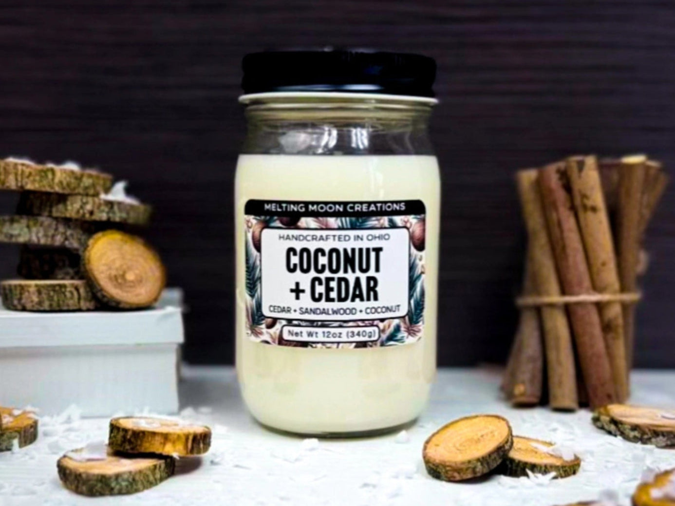 Coconut + Cedar candle in glass jar with black twist on lid sitting on white surface with dark gray background. Wood rounds scattered around with a stack to the left of the candle. A bundle of round wooden sticks to the right. Coconut shavings scattered throughout. Candle label designed by Melting Moon Creations.