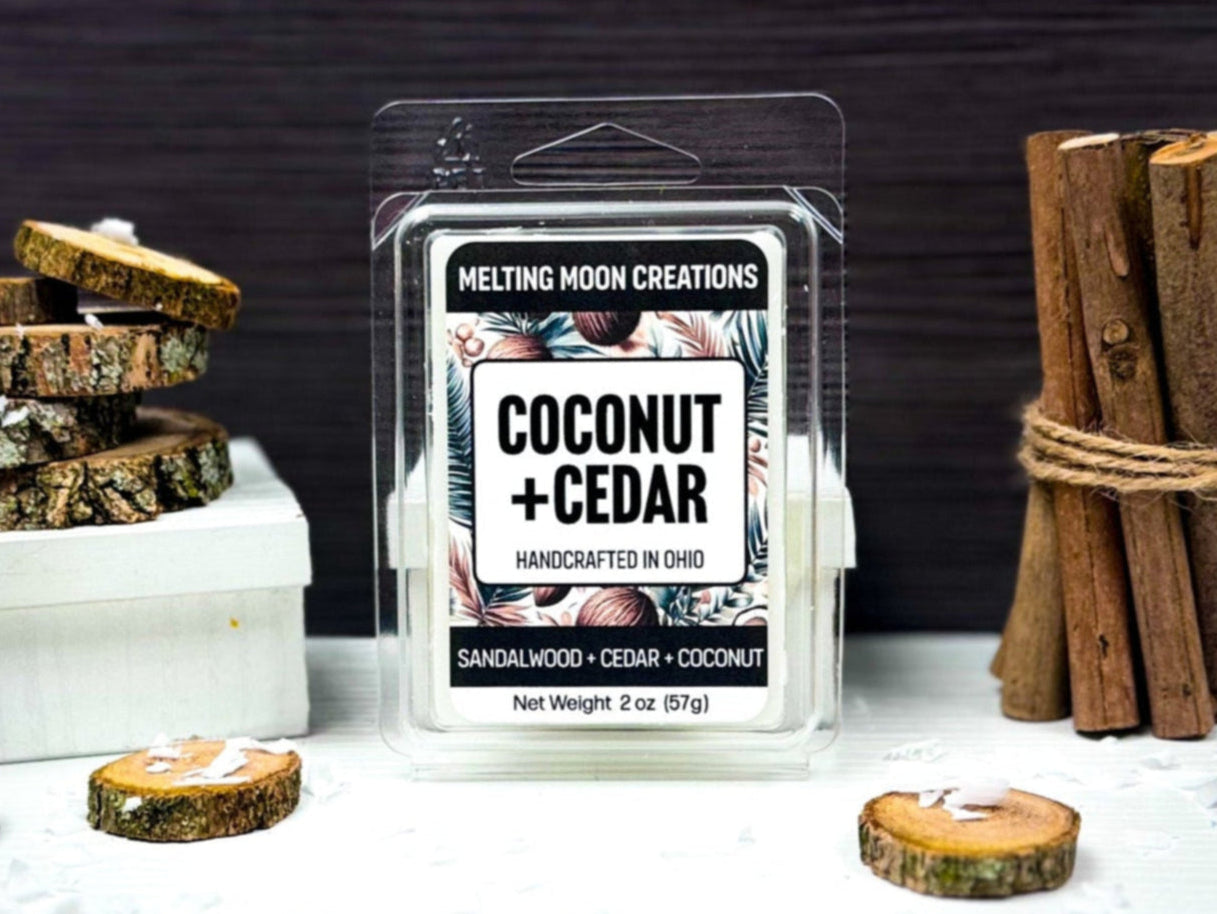 Coconut + Cedar wax melt sitting on white tabletop with dark gray background. Woods rounds scattered around with a stack of five sitting to the left. Bundle of round wood pieces to the right. Coconut shavings sprinkled throughout. Label designed by Melting Moon Creations.