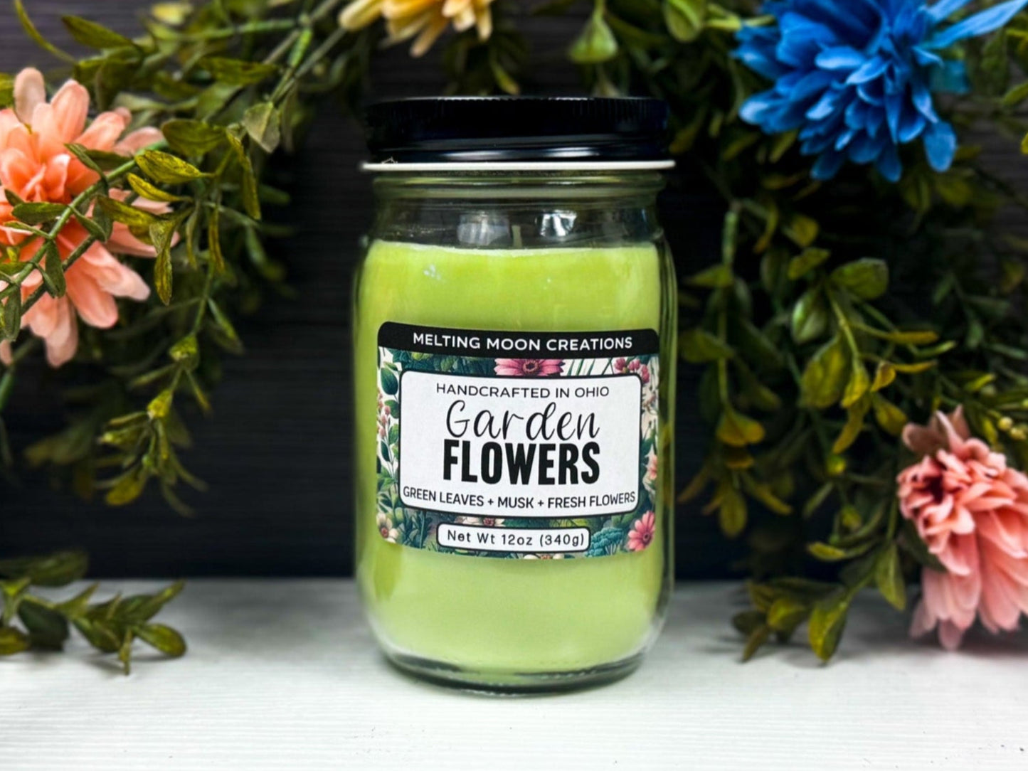 Garden Flowers candle in glass jar with black twist on lid sitting on a white tabletop. Leafy greenery arched behind candle with colorful pops of flowers. Label designed by Melting Moon Creations.