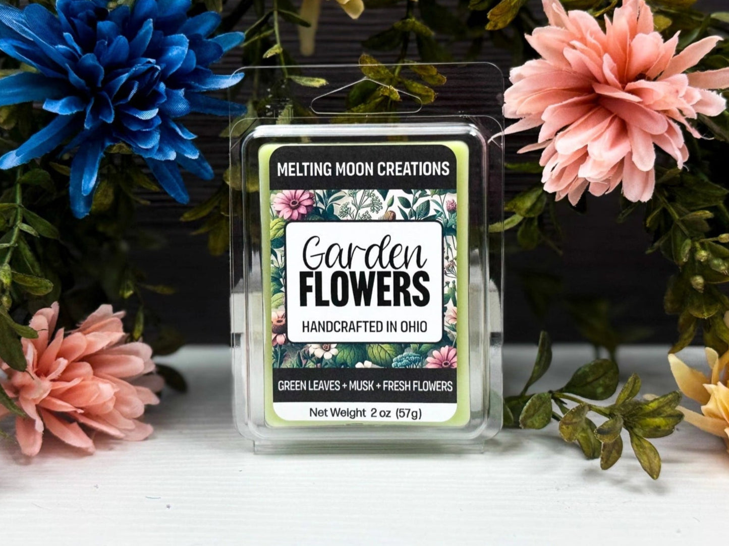 Garden Flowers wax melt sitting on white tabletop. Leafy greenery with colorful flowers arched over melt. Label designed by Melting Moon Creations.
