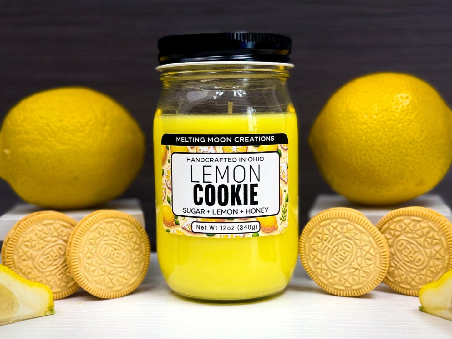Lemon Cookie candle in glass jar with black twist on lid sitting on a white tabletop. A lemon on both sides of wax melt and a lemon slice on the edges. Two lemon-flavored cookies staged on each side. Candle label designed by Melting Moon Creations.