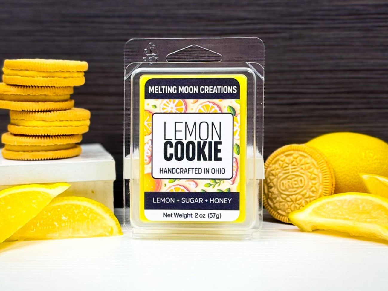 Lemon Cookie wax melt sitting on white counter with dark gray background. Lemons on both sides on wax melt with lemon slices on either side with one lemon-flavored cookie on the right, and a stack of 4 on the left. Labeled by Melting Moon Creations.