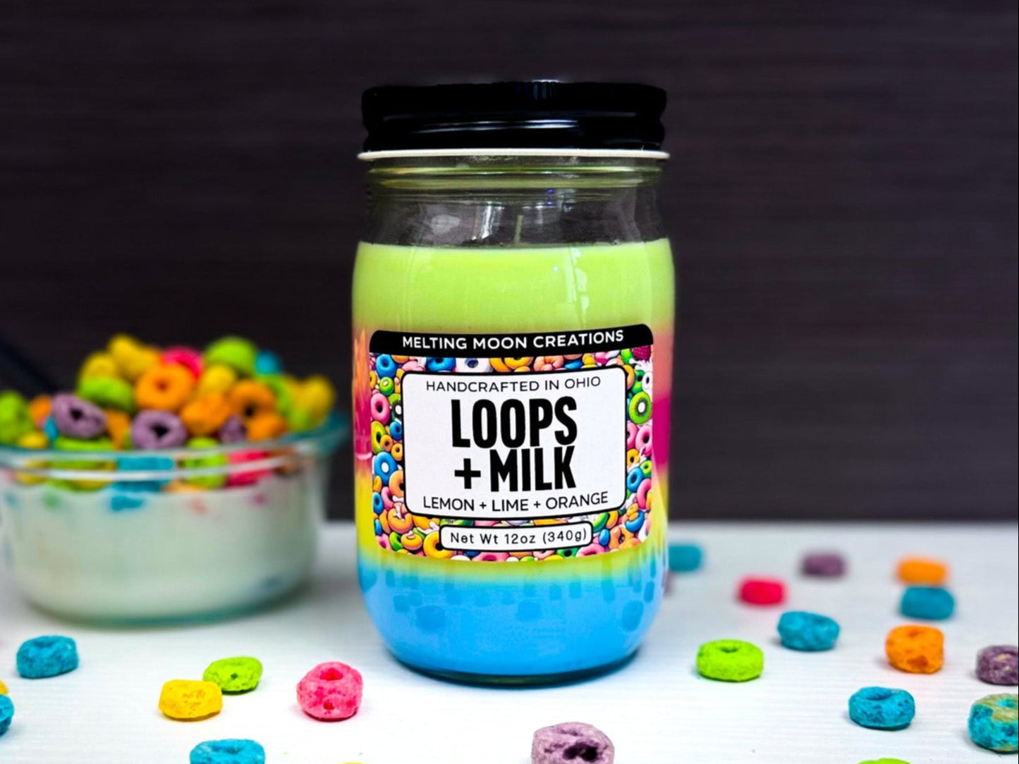 Loops + Milk candle in glass jar with black screw on lid sitting on a white tabletop with dark gray background. Candle wax is layered in five rainbow colors. Small bowl of cereal to the left of candle. Colorful round cereal pieces sprinkled around. Candle label designed by Melting Moon Creations.