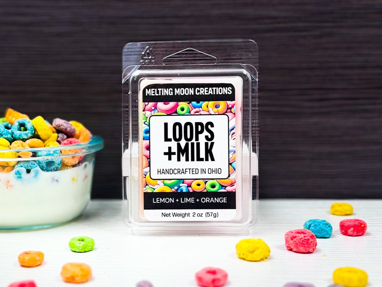 Loops + Milk wax melt sitting on white surface with dark gray background. Small bowl of cereal to left of melt, colorful cereal pieces scattered across tabletop.