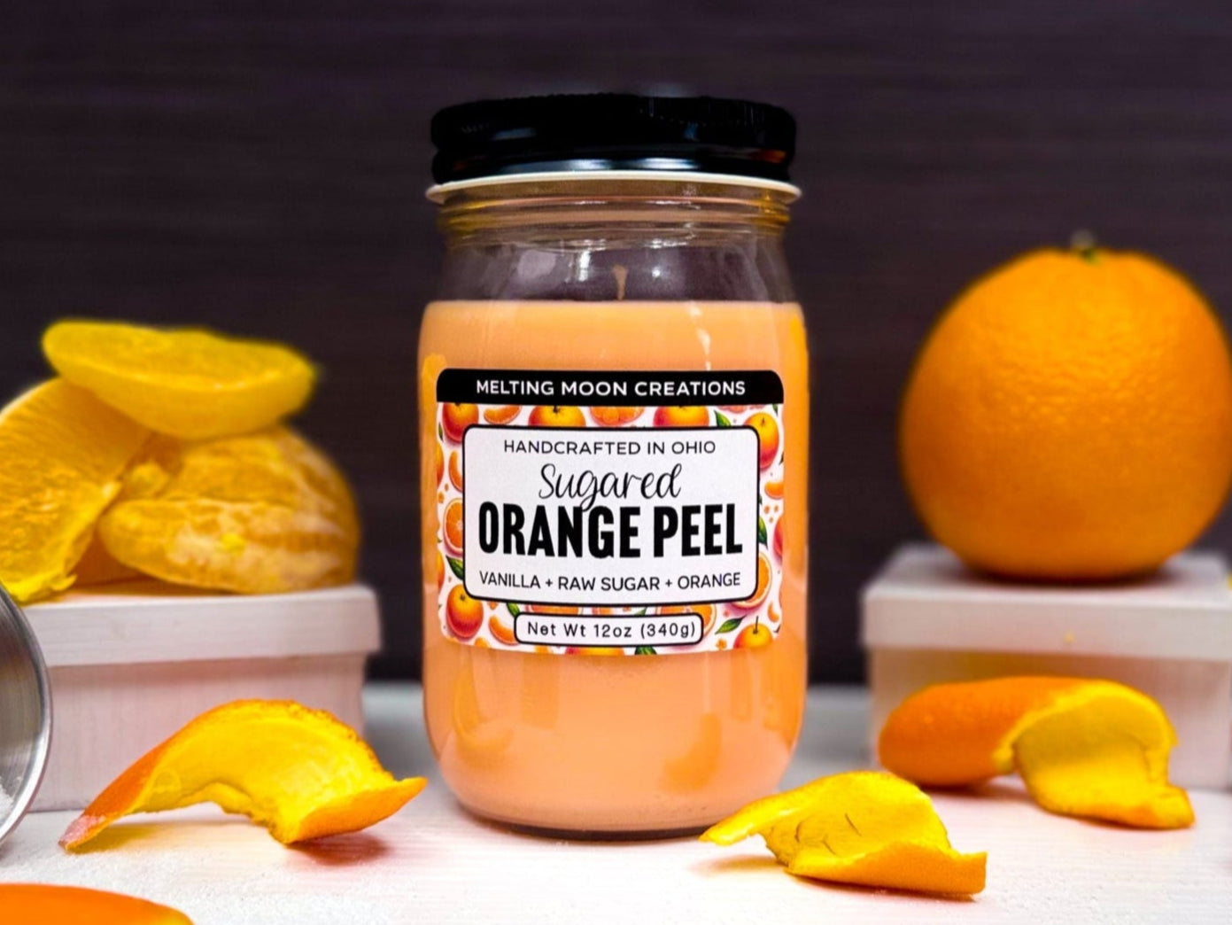 Sugared Orange Peel candle in glass jar with black screw on lid sitting on a white surface with a dark gray background. Orange to the right, and orange slices and peels scattered around. Container of sugar tipped over in far left. Label designed by Melting Moon Creations.