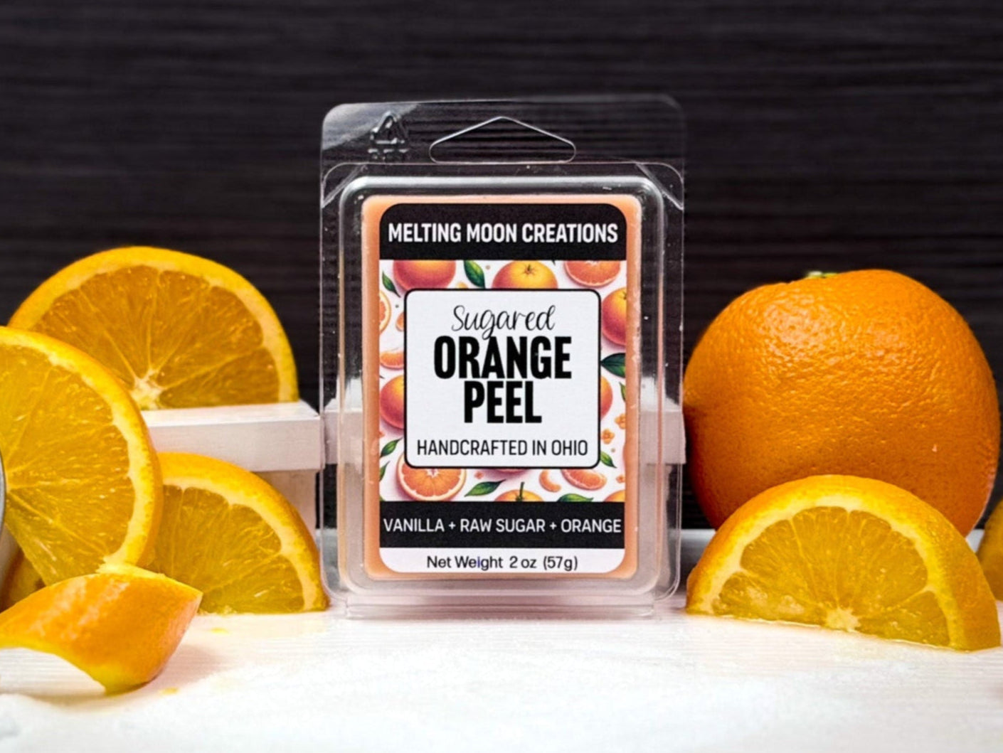 Sugared Orange Peel wax melt sitting on white surface with dark gray background. Orange sitting to the right with orange slices on both sides. Container of sugar sitting to far left, tipped over. Label designed by Melting Moon Creations.