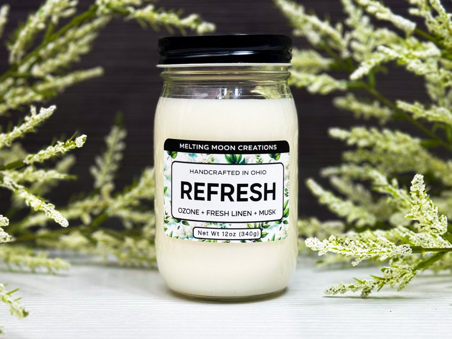Refresh candle in a glass jar with black screw on lid. Green and white leafy props in background. Candle label designed by Melting Moon Creations.