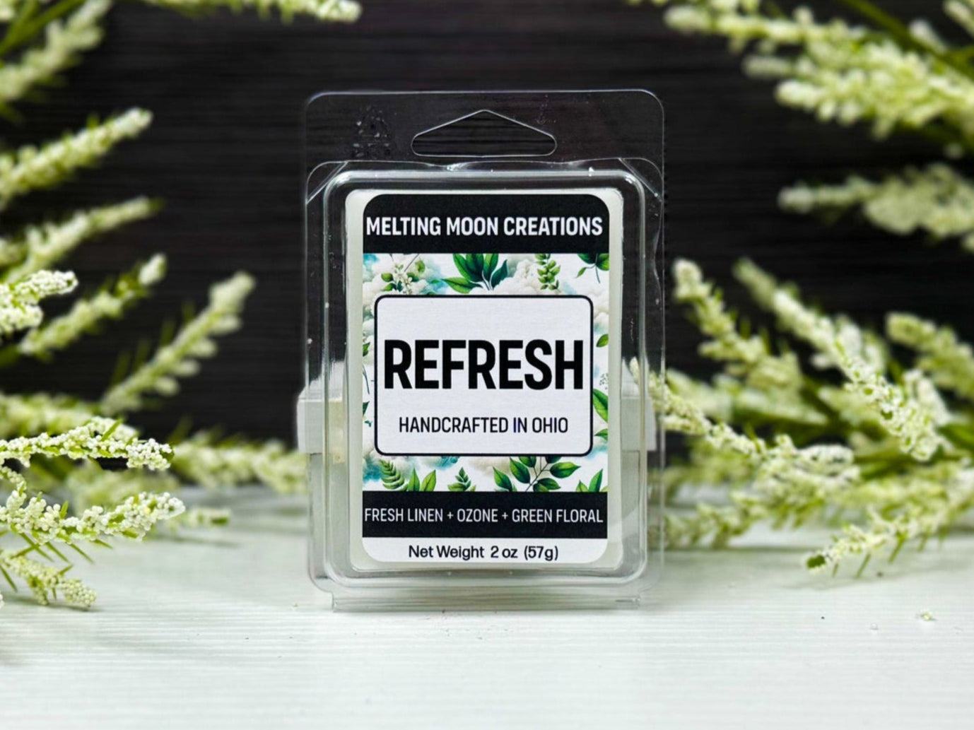 Refresh wax melt sitting on white tabletop with dark gray background. Green and white leafy plants staged behind. Wax melt label designed by Melting Moon Creations.