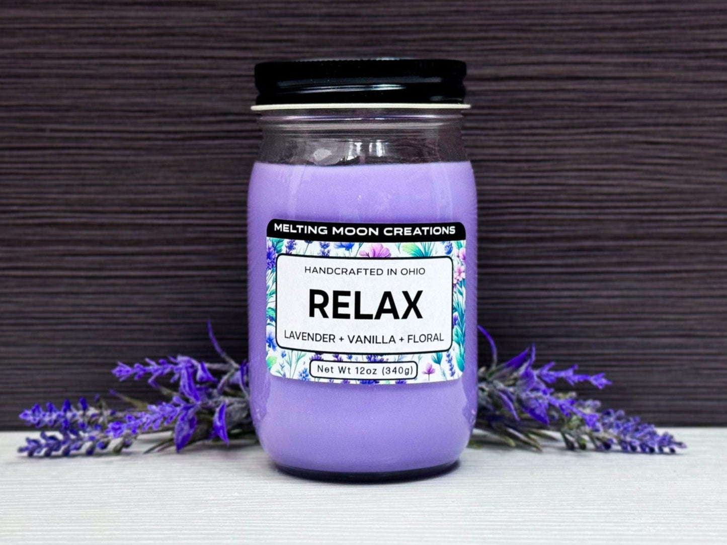 Relax candle in glass jar with black twist on lid sitting on white tabletop with dark gray background. Lavender flowers on both sides on the candle. Candle wax is purple. Label designed by Melting Moon Creations.