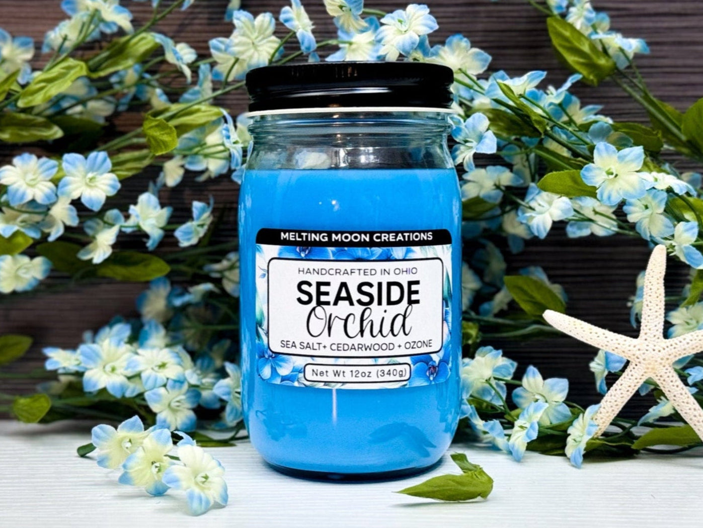 Seaside Orchid candle in a glass jar with black twist on lid sitting on a white tabletop with a dark gray background. Blue and white flowers placed behind candle with a dried starfish to the right. Label designed by Melting Moon Creations.