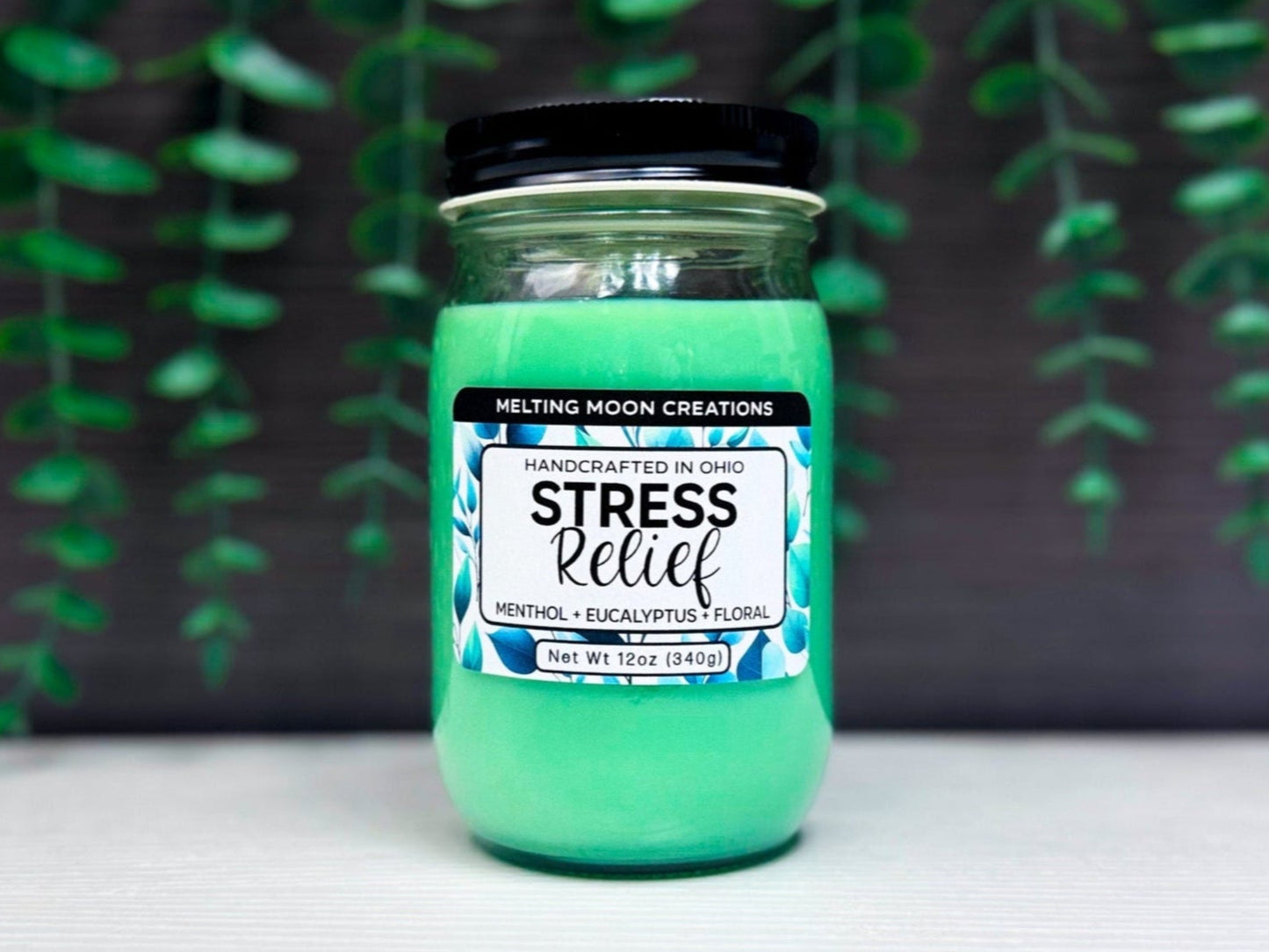 Stress Relief candle in glass jar with black screw on lid sitting on a white tabletop with dark gray background with leaves hanging down. Green candle wax. Label designed by Melting Moon Creations.