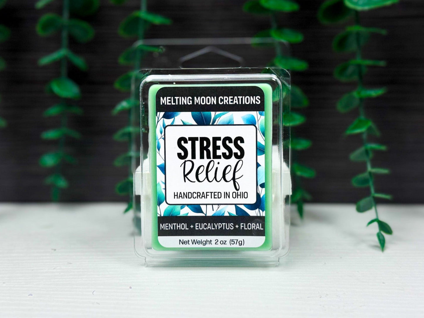 Stress Relief wax melt sitting on white tabletop with dark gray background. Green leaves hanging behind wax melt. Label designed by Melting Moon Creations.