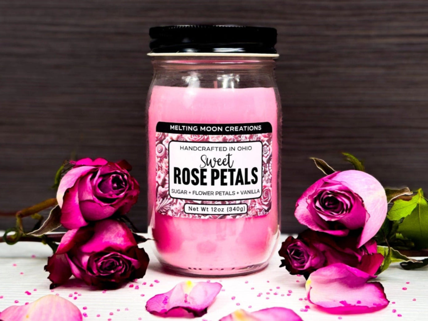 Sweet Rose Petals in glass jar with black twist on lid on white tabletop with dark gray background. 4 roses total with two stacked on either side of candle with rose petals and pink sugar crystals sprinkled around.