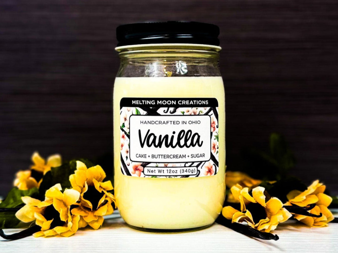 Cream white vanilla candle sitting on white surface with dark gray background. Vanilla beans and flowers laying on surface. Label designed by Melting Moon Creations.