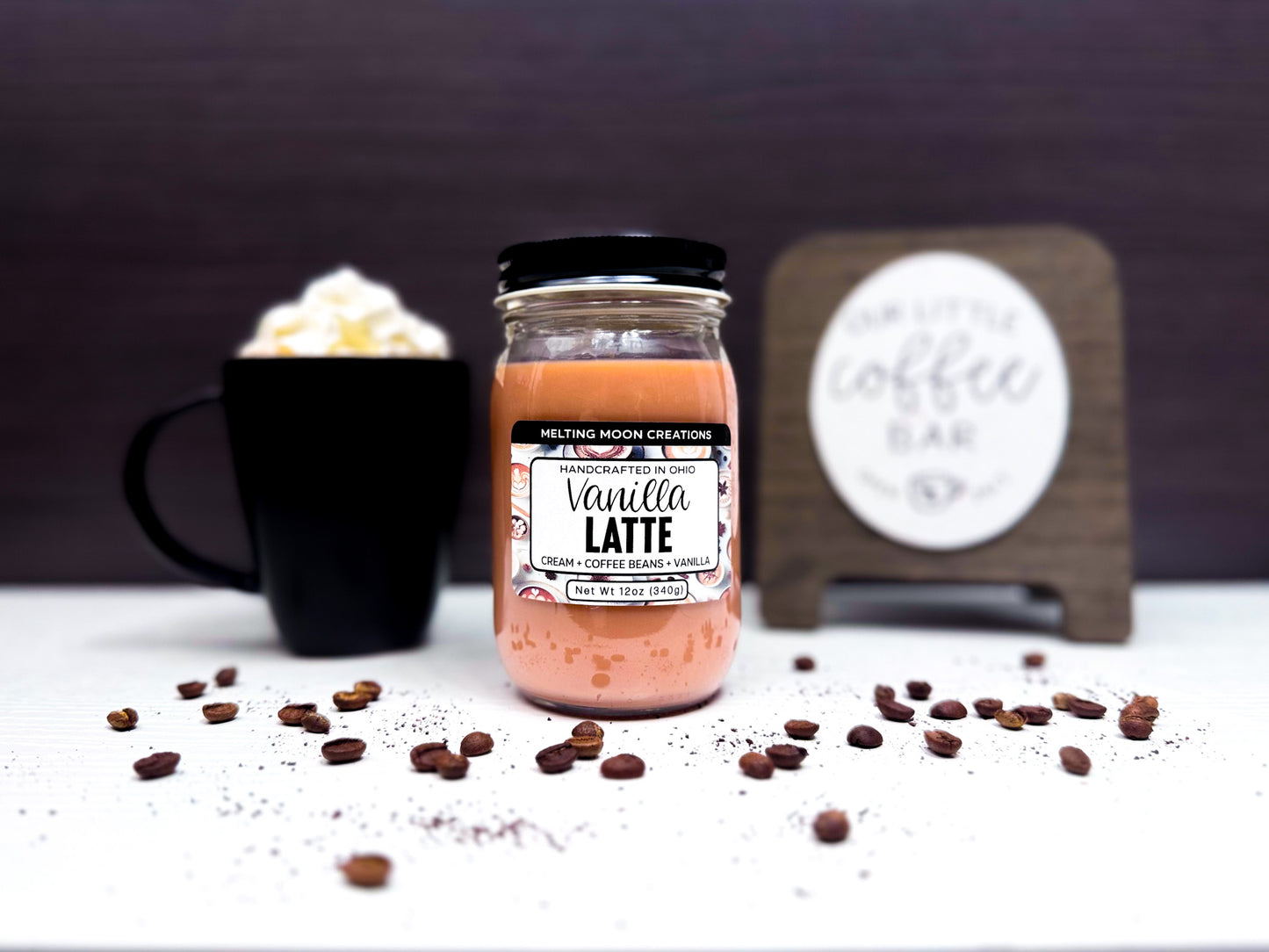 Vanilla Latte candle in glass jar with black screw on lid sitting on a white surface with a dark gray background. A coffee bar sign and a coffee drink in a black mug with whipped cream are staged behind it. Label designed by Melting Moon Creations.