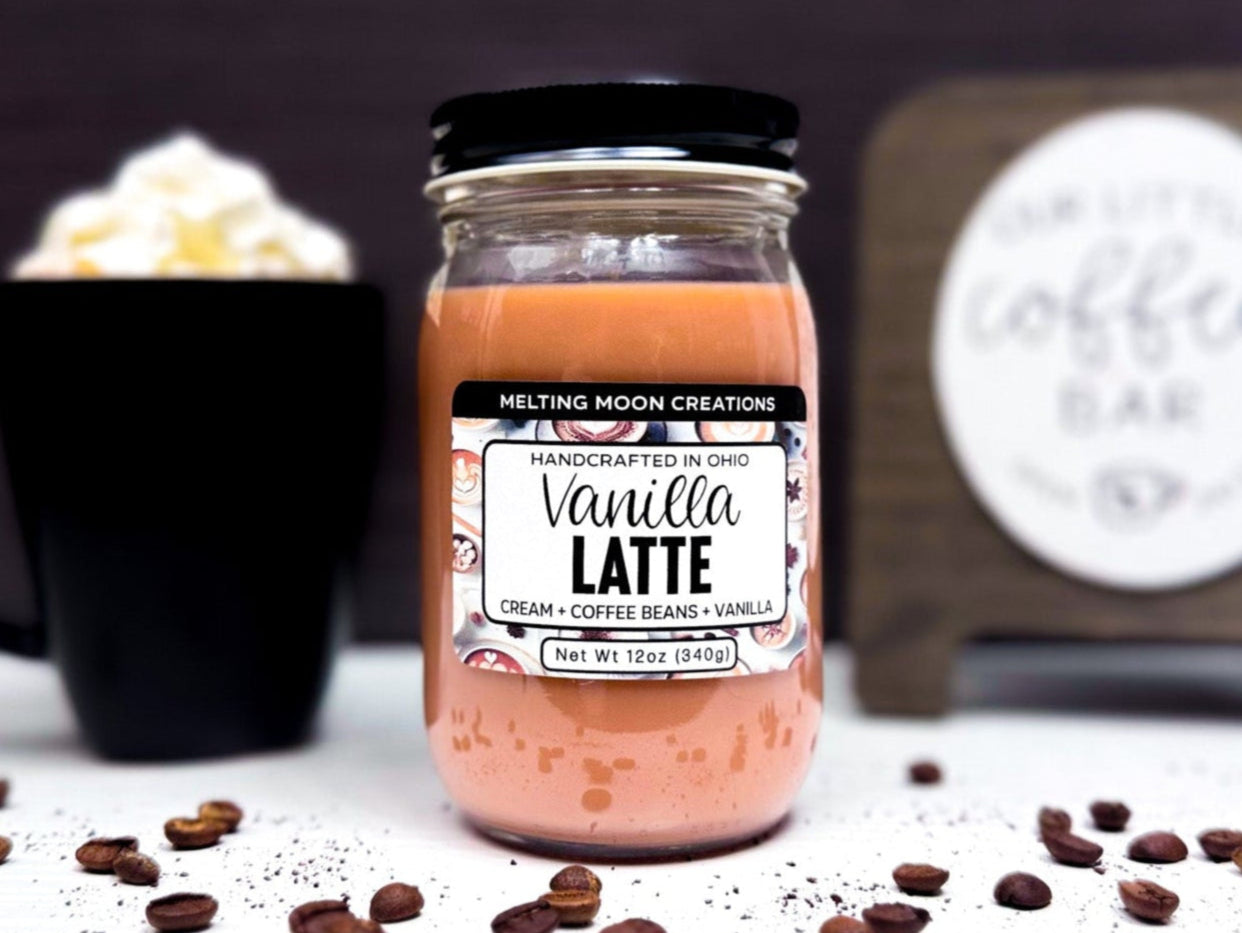 Vanilla Latte candle in glass jar with black screw on lid sitting on a white surface with a dark gray background. A coffee bar sign and a coffee drink in a black mug with whipped cream are staged behind it. Label designed by Melting Moon Creations.