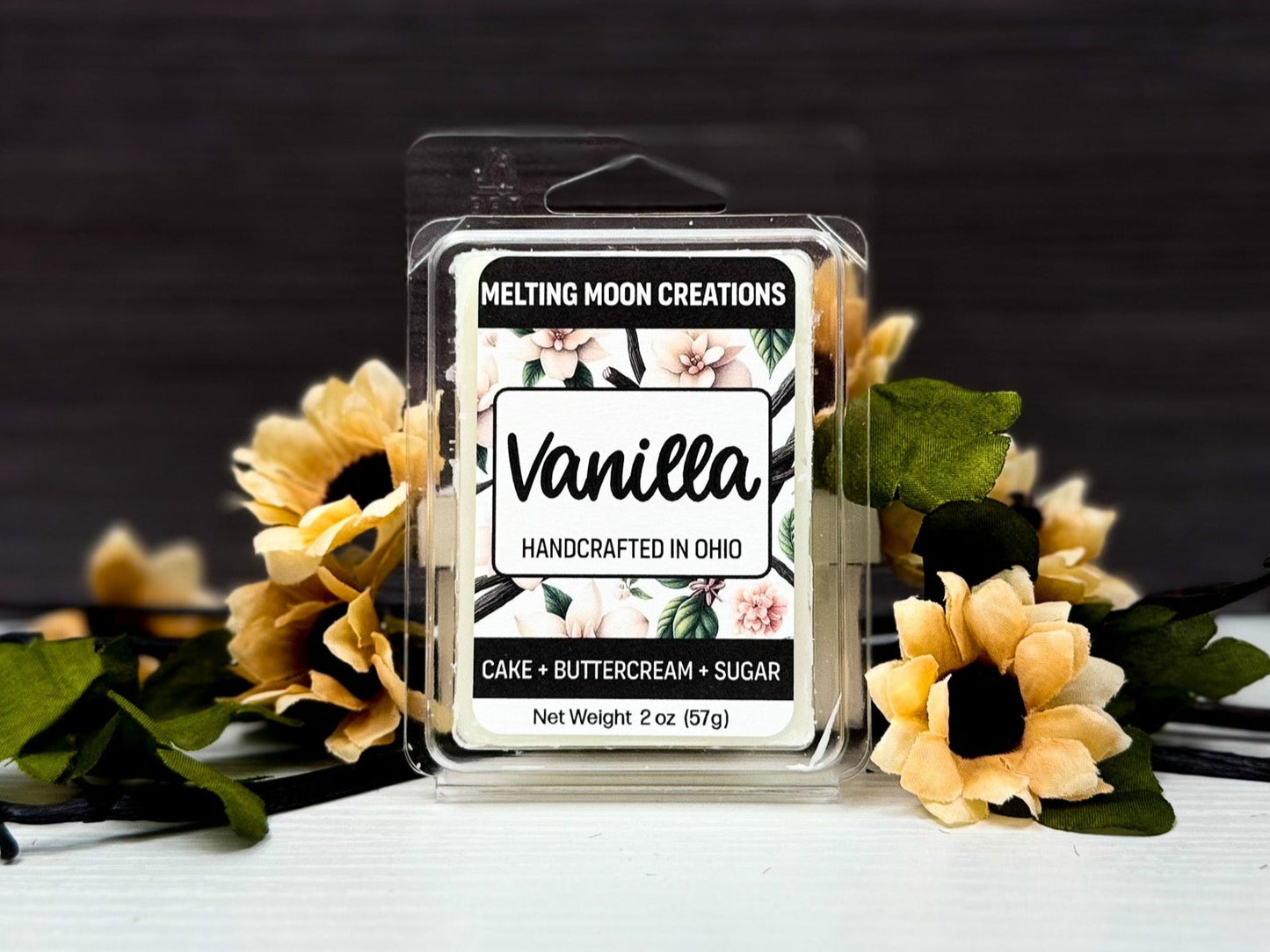 Vanilla wax melt sitting on white surface with dark gray background. Vanilla beans and cream colored flowers staged around. Label designed by Melting Moon Creations.