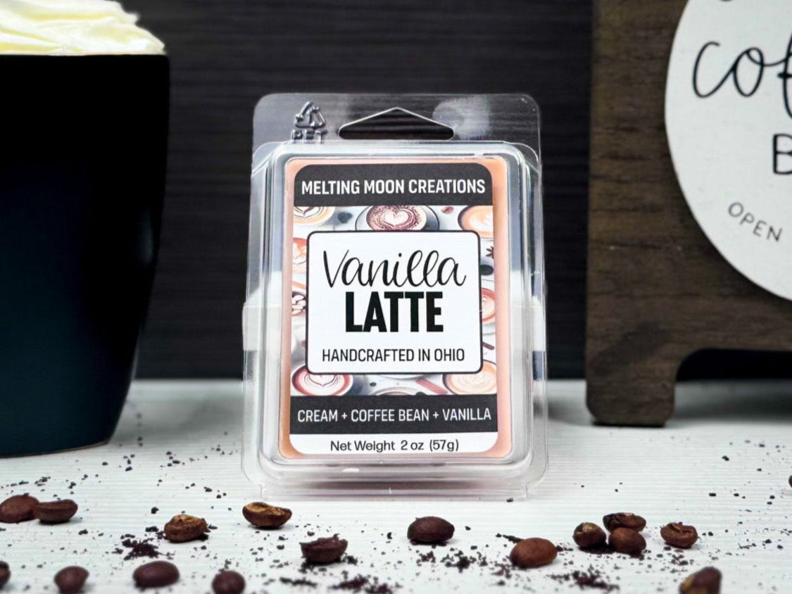 Vanilla Latte wax melt on white surface with dark gray background. Black mug on left with a latte with whipped cream, and a wooden coffee bar sign to the right. Coffee beans scattered across surface.