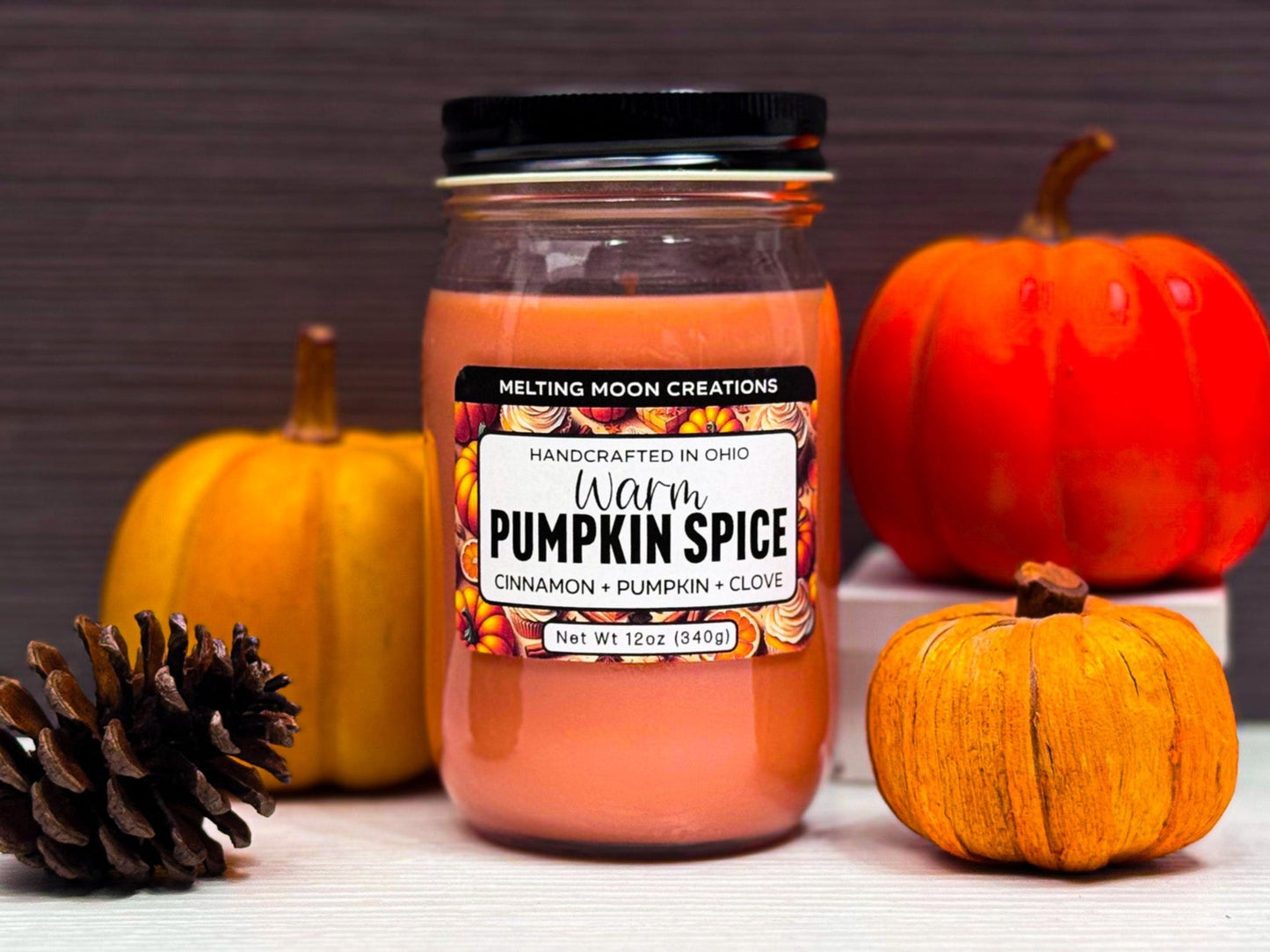 Warm Pumpkin Spice candle in glass jar with black twist on lid on a white surface with dark gray background. Surrounded by small pumpkins and a pinecone. Label designed by Melting Moon Creations.