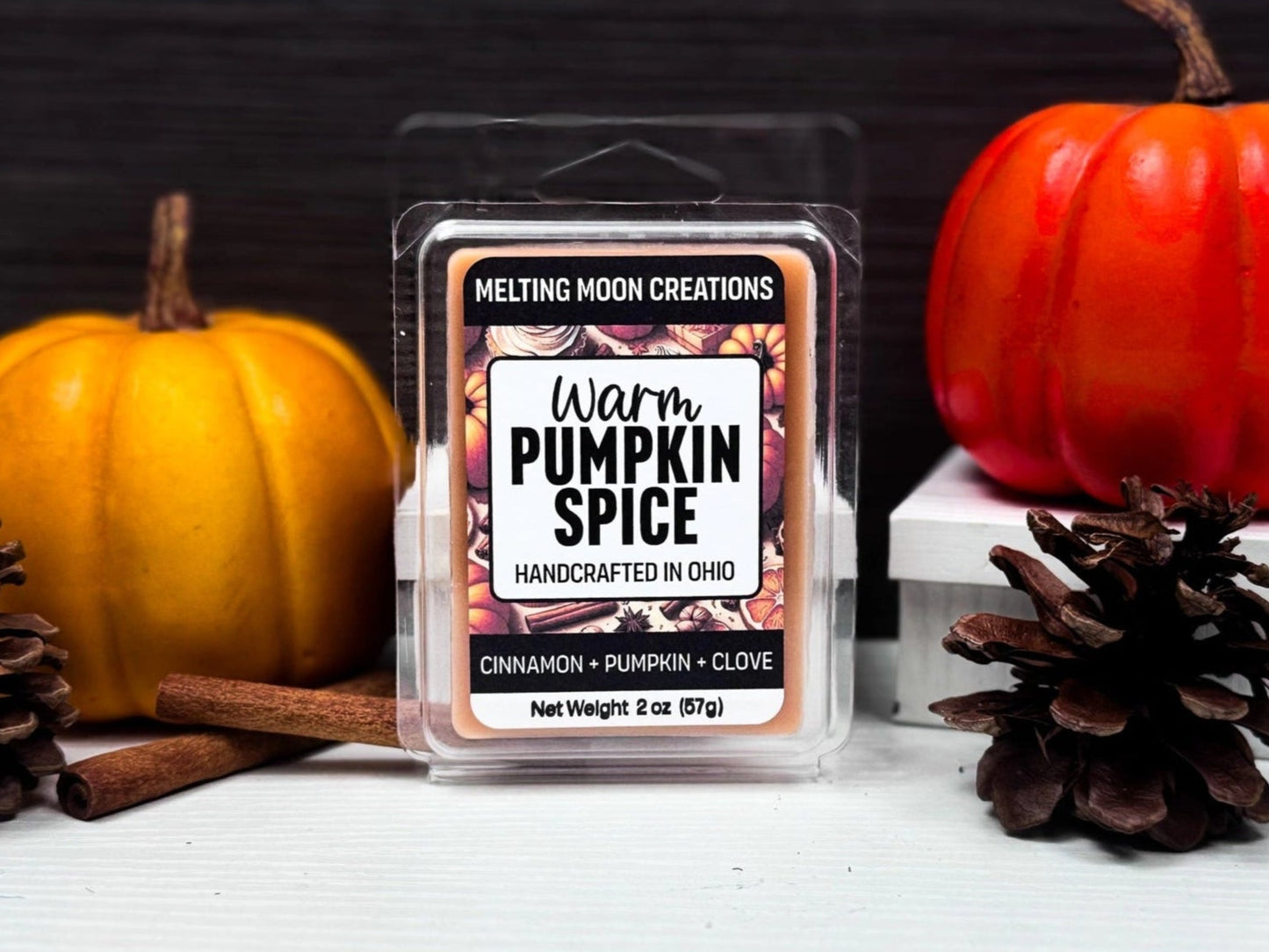 Warm Pumpkin Spice wax melt sitting on white tabletop with dark gray background. Large bright pumpkin to the right and a light orange pumpkin to the left. One pine cone on each side and two cinnamon sticks on the left. Label designed by Melting Moon.