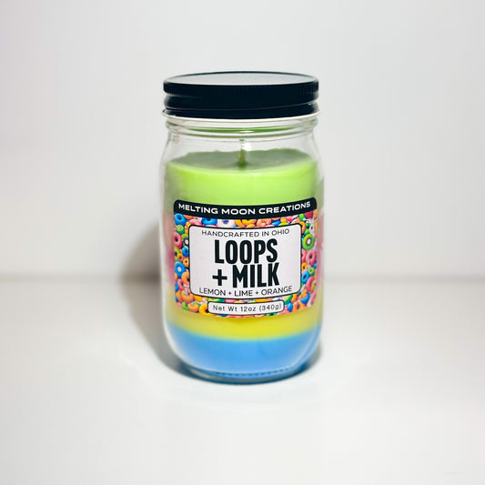 Loops + Milk candle in a glass jar with black twist on lid sitting on a white backdrop. Candle wax is dyed in even layers of rainbow colors. Candle label designed by Melting Moon Creations.