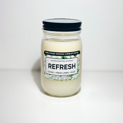 Refresh candle in glass jar with black screw on lid sitting on white backdrop. Candle label designed by Melting Moon Creations.