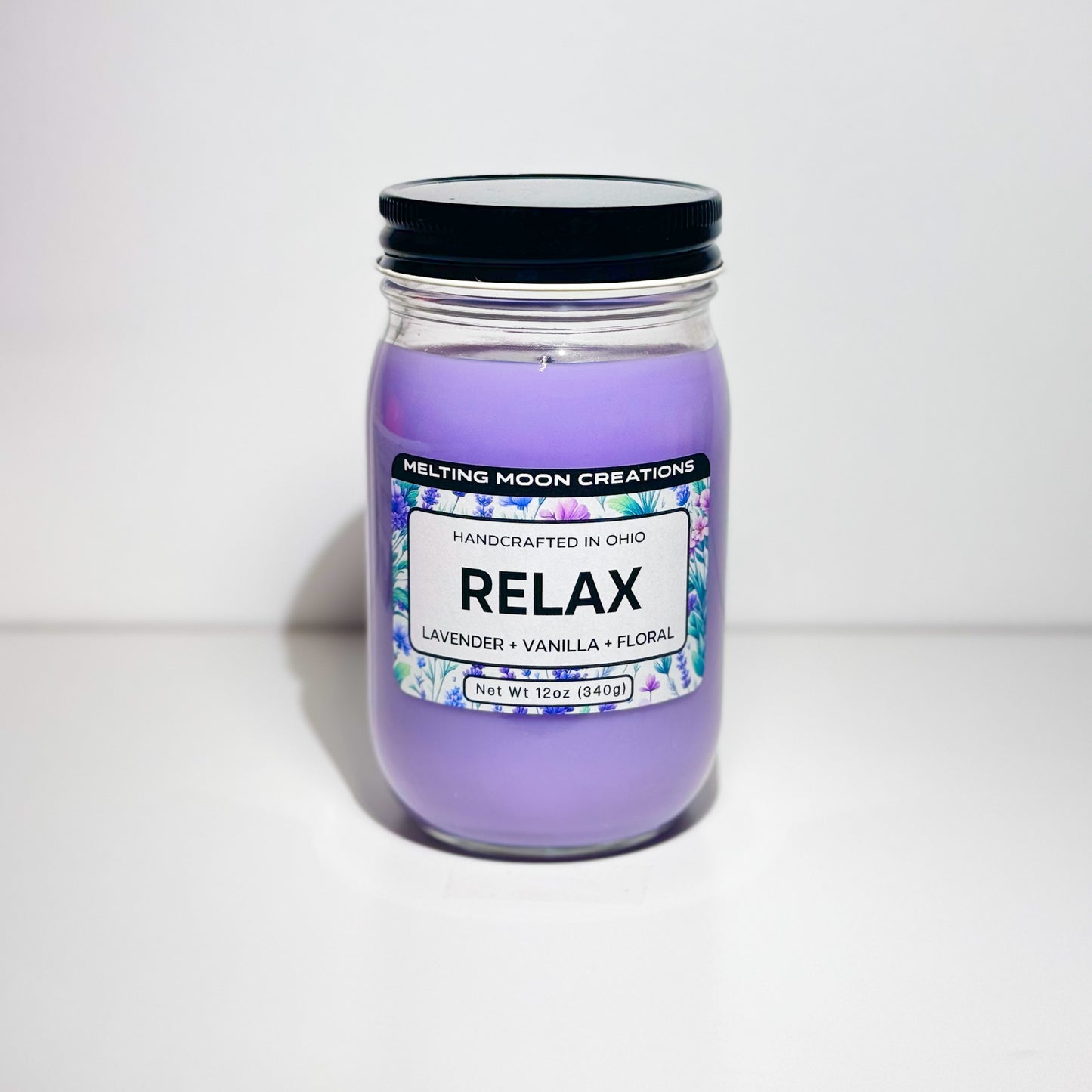 Relax candle in glass jar with black twist on lid sitting on white backdrop. Candle wax is purple. Candle label designed by Melting Moon Creations.