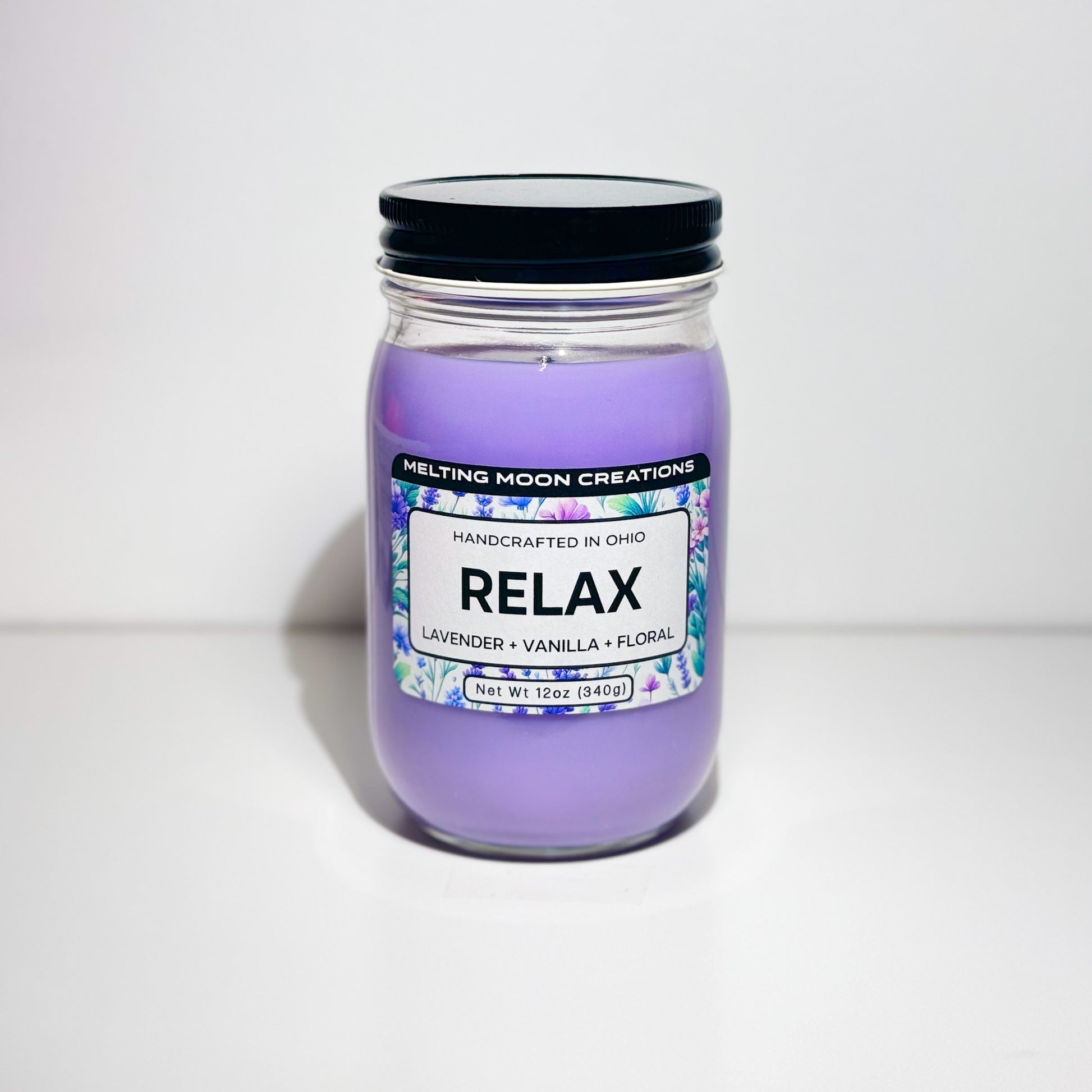 Relax candle in glass jar with black twist on lid sitting on white backdrop. Candle wax is purple. Candle label designed by Melting Moon Creations.