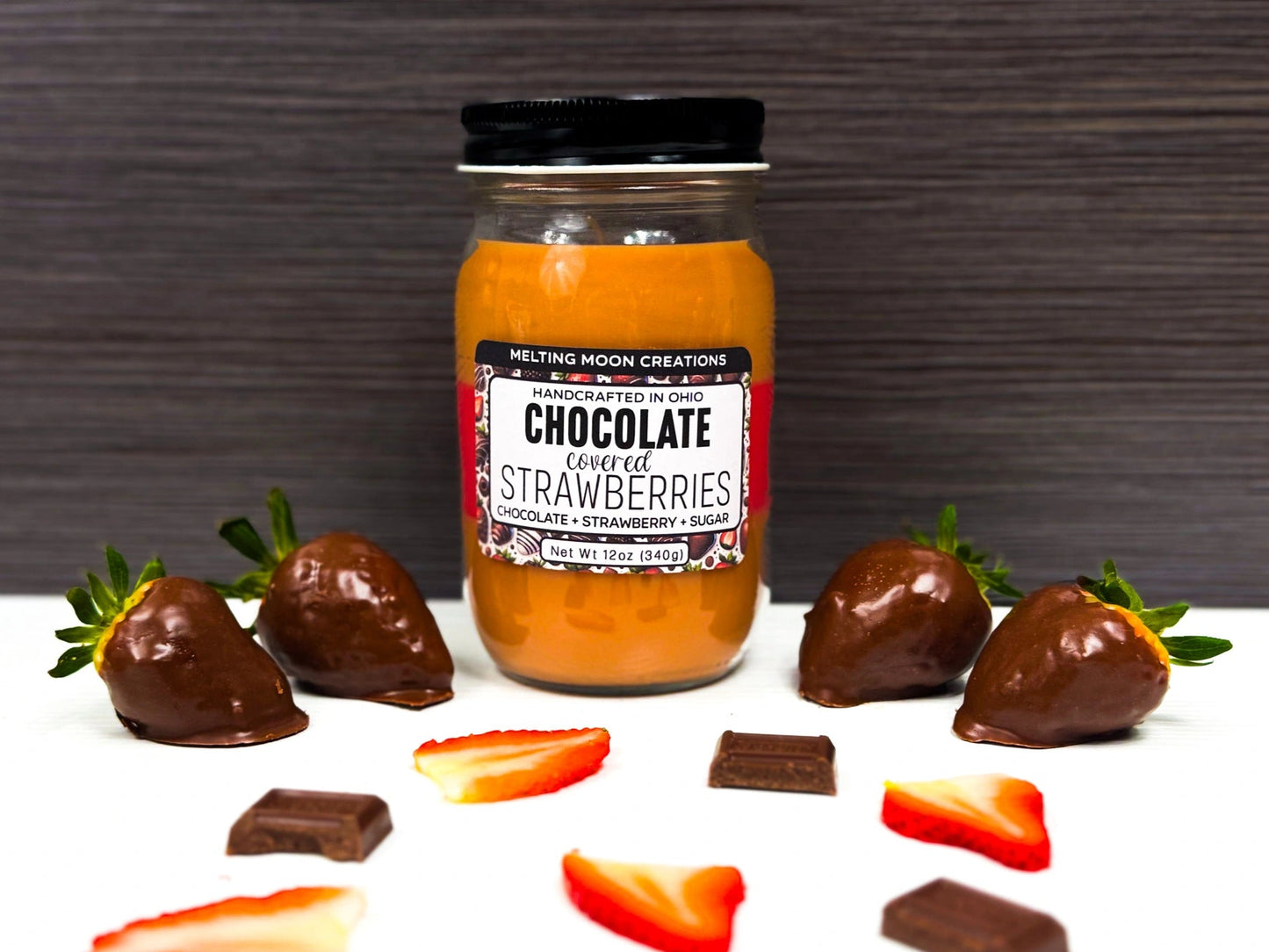 Chocolate Covered Strawberries candle in glass jar sitting on a white surface with a dark gray background. 2 chocolate strawberries on each side. Slices of strawberries and pieces of chocolate laying around.