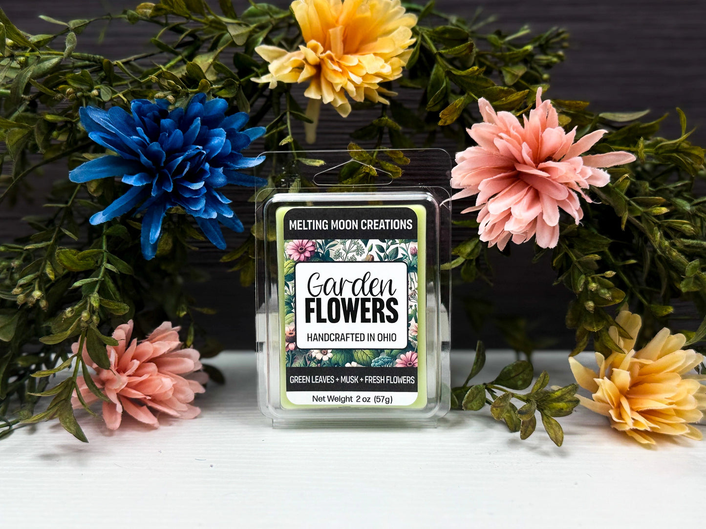 Garden Flowers wax melt sitting on white tabletop. Leafy greenery with colorful flowers arched over melt. Label designed by Melting Moon Creations.