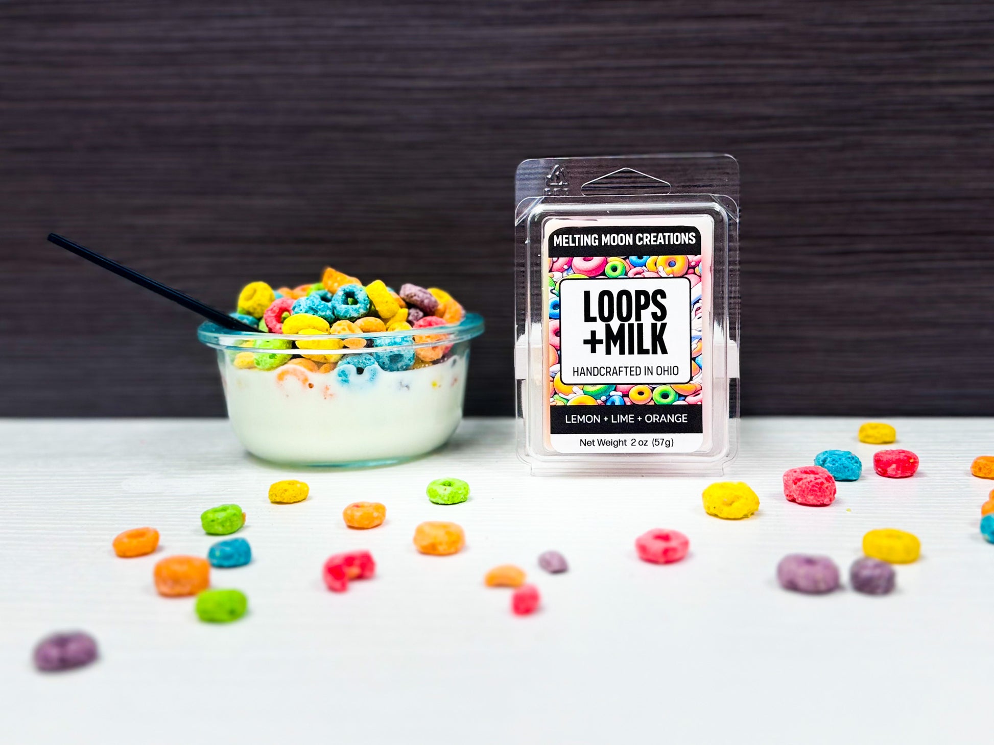 Loops + Milk wax melt sitting on white surface with dark gray background. Small bowl of cereal to left of melt, colorful cereal pieces scattered across tabletop.