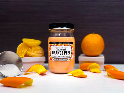 Sugared Orange Peel candle in glass jar with black screw on lid sitting on a white surface with a dark gray background. Orange to the right, and orange slices and peels scattered around. Container of sugar tipped over in far left. Label designed by Melting Moon Creations.