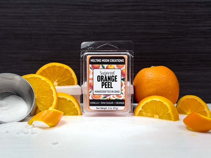 Sugared Orange Peel wax melt sitting on white surface with dark gray background. Orange sitting to the right with orange slices on both sides. Container of sugar sitting to far left, tipped over. Label designed by Melting Moon Creations.