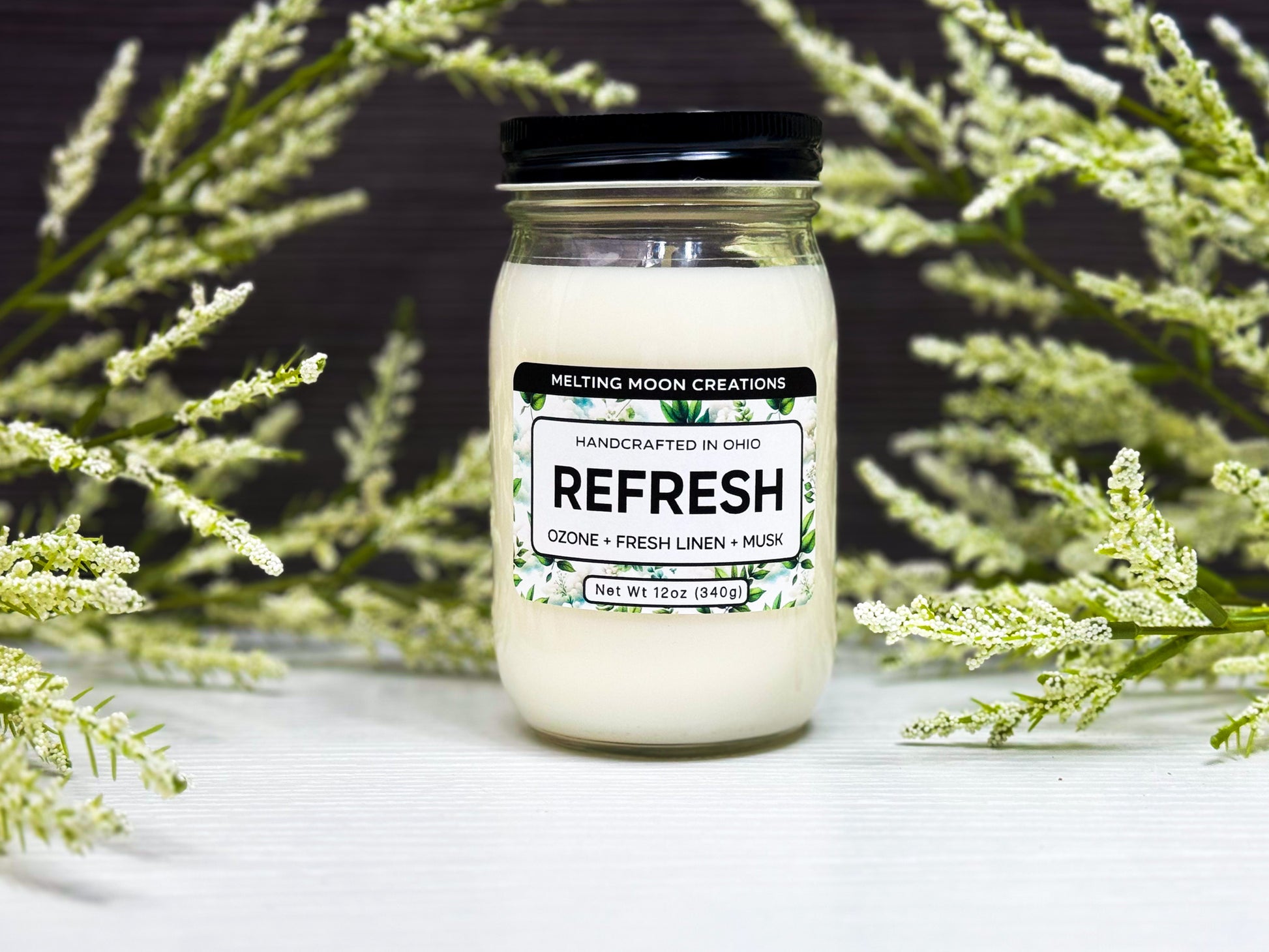 Refresh candle in a glass jar with black screw on lid. Green and white leafy props in background. Candle label designed by Melting Moon Creations. 