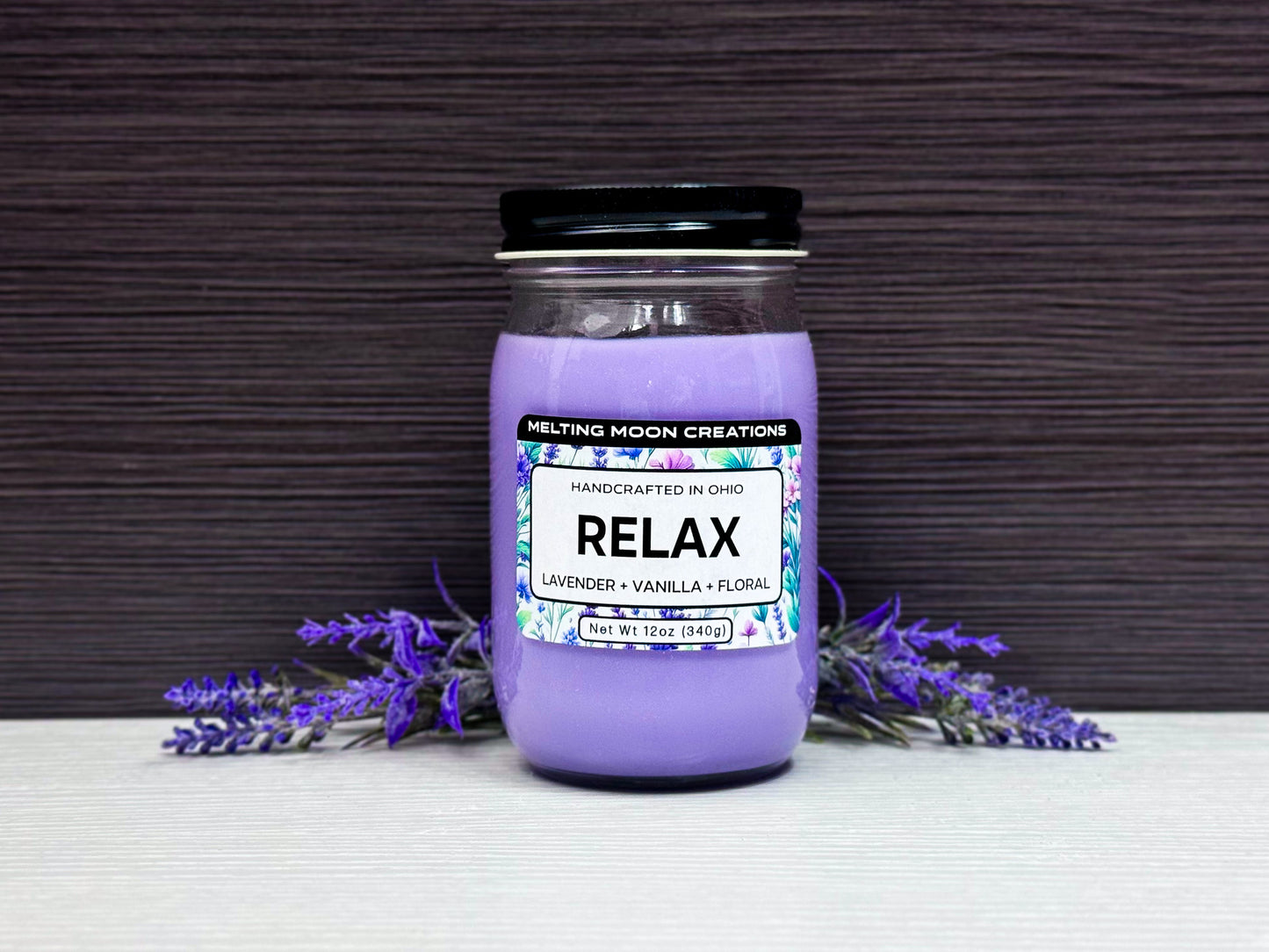 Relax candle in glass jar with black twist on lid sitting on white tabletop with dark gray background. Lavender flowers on both sides on the candle. Candle wax is purple. Label designed by Melting Moon Creations.