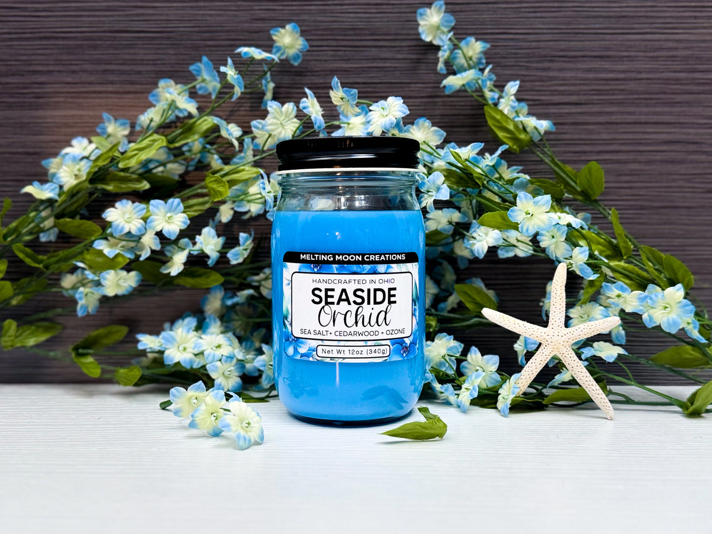 Seaside Orchid candle in a glass jar with black twist on lid sitting on a white tabletop with a dark gray background. Blue and white flowers placed behind candle with a dried starfish to the right. Label designed by Melting Moon Creations.