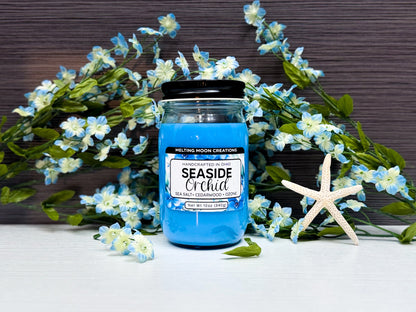 Seaside Orchid candle in a glass jar with black twist on lid sitting on a white tabletop with a dark gray background. Blue and white flowers placed behind candle with a dried starfish to the right. Label designed by Melting Moon Creations.