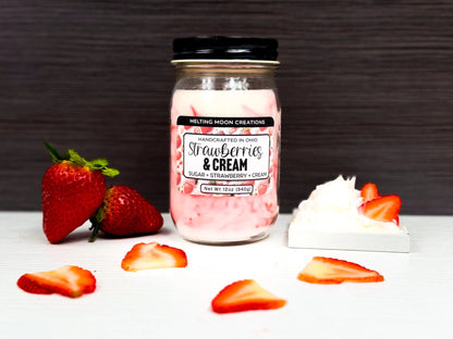 Strawberries and Cream candle in glass jar with black twist on lid sitting on white tabletop with dark gray background. Whipped cream with strawberry slices to the right. 2 whole strawberries staged to the left. Strawberry slices laying around. Candle label designed by Melting Moon.