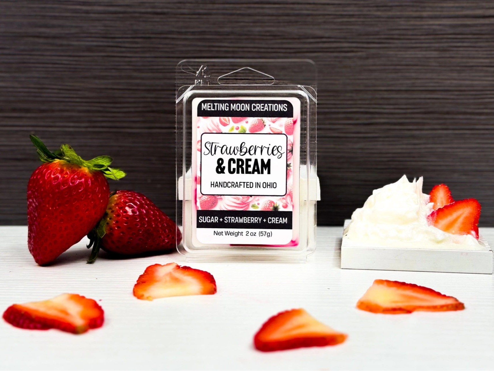 Strawberries and Cream wax melt on white tabletop with dark gray background. Whipped cream with strawberry slices to the right and two whole strawberries stacked to left. Strawberry slices laying around.