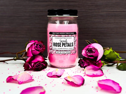 Sweet Rose Petals in glass jar with black twist on lid on white tabletop with dark gray background. 4 roses total with two stacked on either side of candle with rose petals and pink sugar crystals sprinkled around.