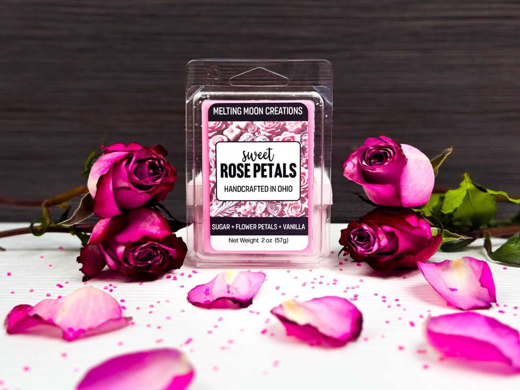 Sweet Rose Petals wax melt on white tabletop with dark gray background. 4 roses in total with 2 stacked on each side of the wax melt. Rose petals and pink sugar crystals sprinkled around.