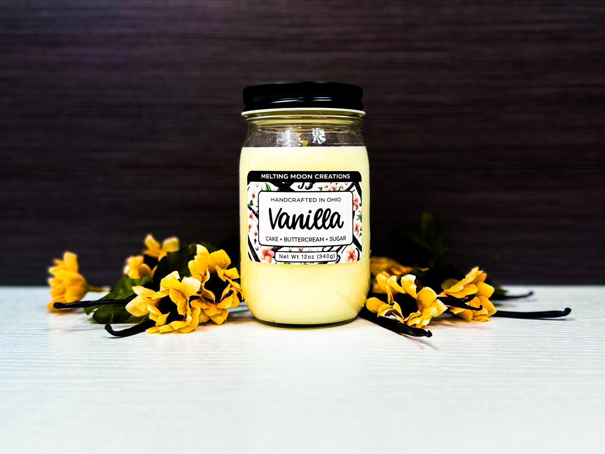 Cream white vanilla candle sitting on white surface with dark gray background. Vanilla beans and flowers laying on surface. Label designed by Melting Moon Creations.