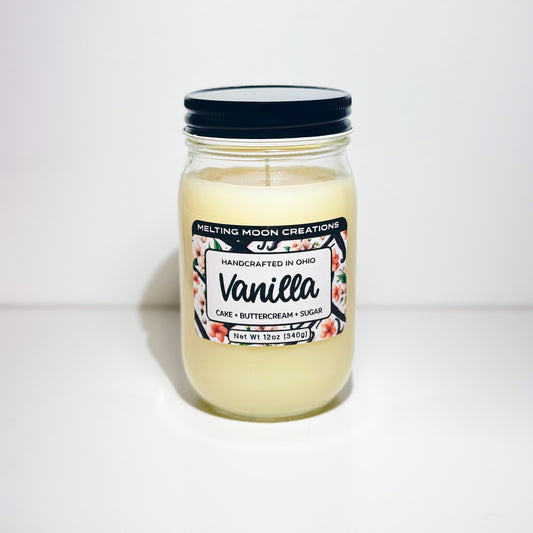 Cream white Vanilla scented candle in a glass jar with black screw on lid. Sitting on white backdrop. Label designed by Melting Moon Creations.