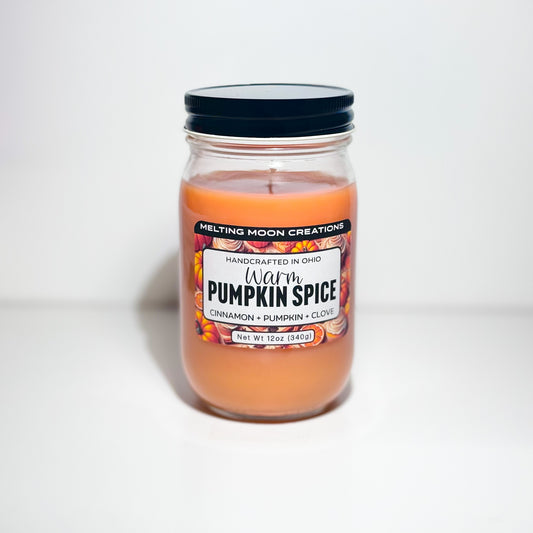 Warm pumpkin spice dyed candle in a glass jar with black screw on lid sitting on a white backdrop. Label designed by Melting Moon Creations. 